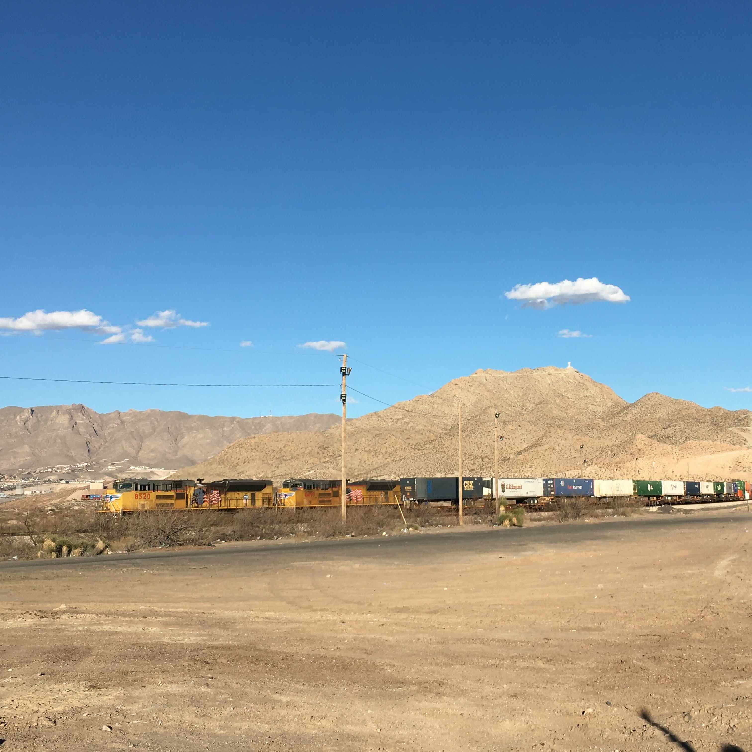 Panoramic photo of a train running over a desert landscape, blue sky overhead.